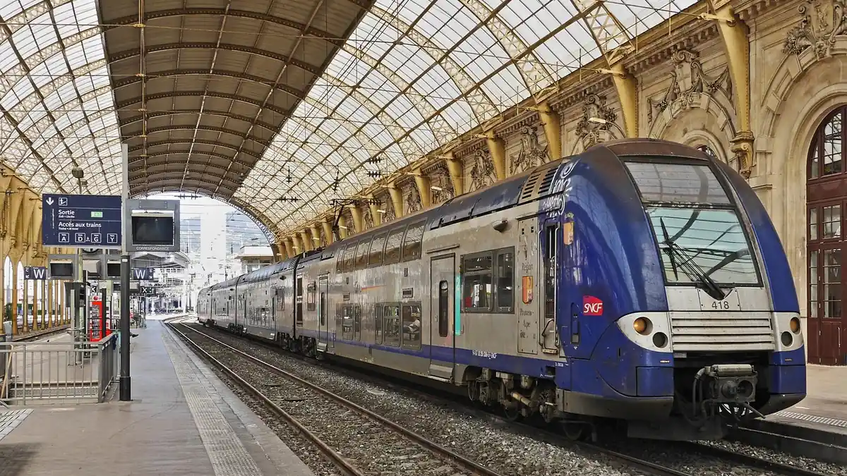 Rame SNCF bleue à deux niveaux arrêtée à quai sous la grande verrière métallique d'une gare, panneau de sortie devant
