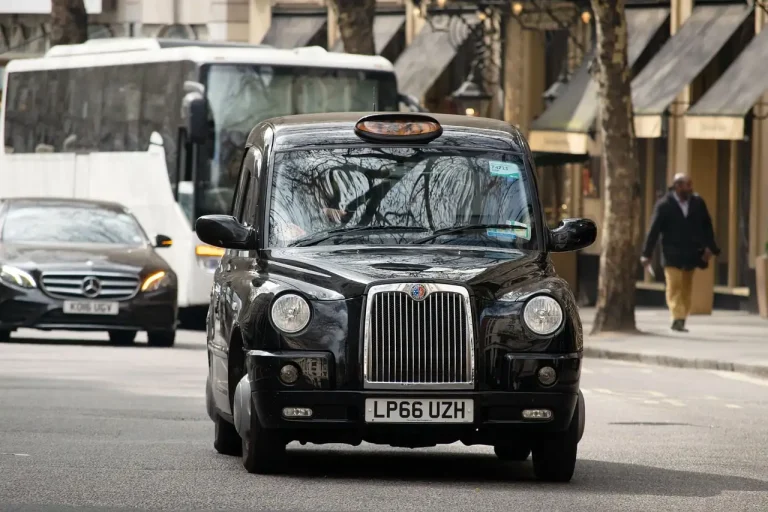 Taxi noir londonien vu de face dans une rue commerçante, une berline et un autocar derrière lui