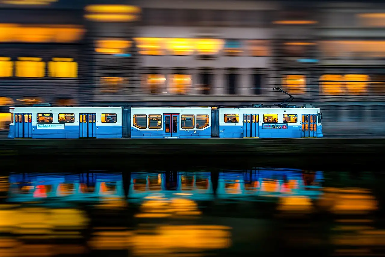 Rame de tramway bleu et blanc en mouvement le soir, fenêtres éclairées reflétées dans le canal