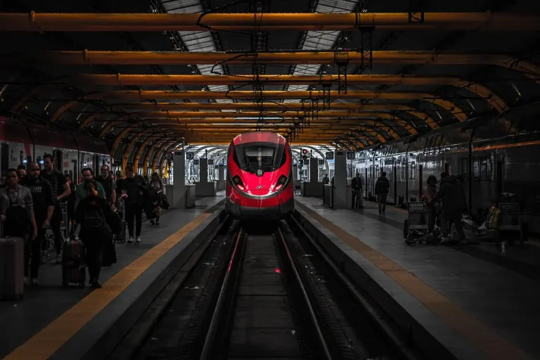 Train rouge à grande vitesse arrêté à quai sous une verrière sombre, voyageurs et valises sur le quai