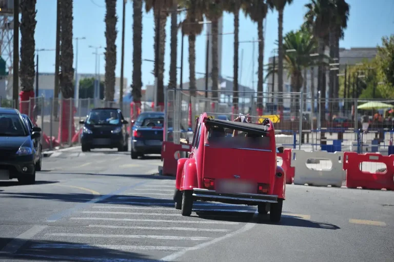 Citroën 2CV rouge décapotée roulant sur une avenue bordée de palmiers, barrières de chantier rouges et blanches