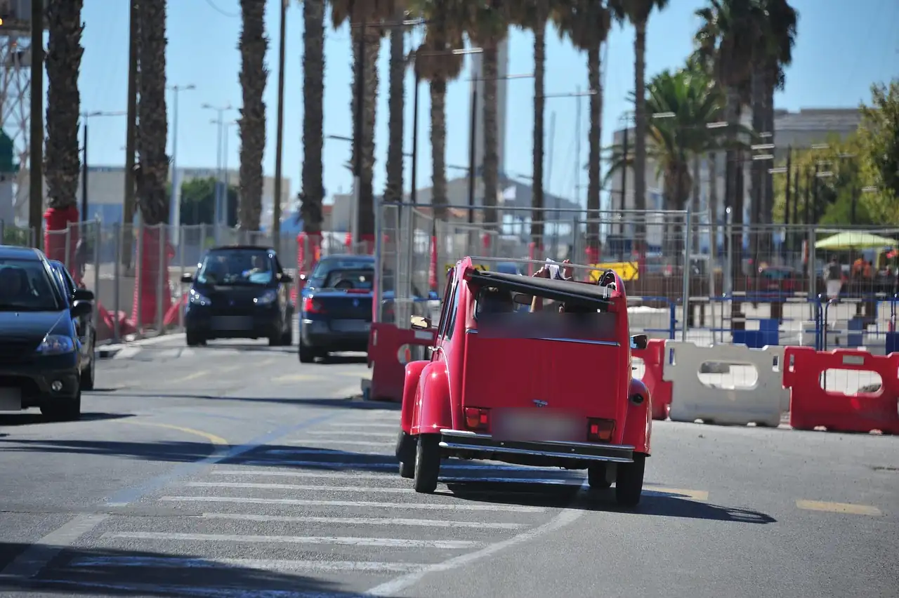 Citroën 2CV rouge décapotée roulant sur une avenue bordée de palmiers, barrières de chantier rouges et blanches