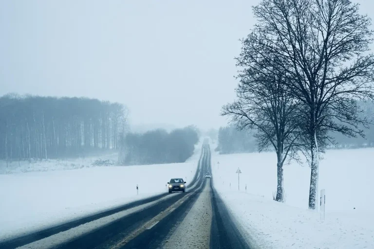 Route de campagne enneigée sous un ciel gris, une voiture phares allumés roule entre les champs blancs