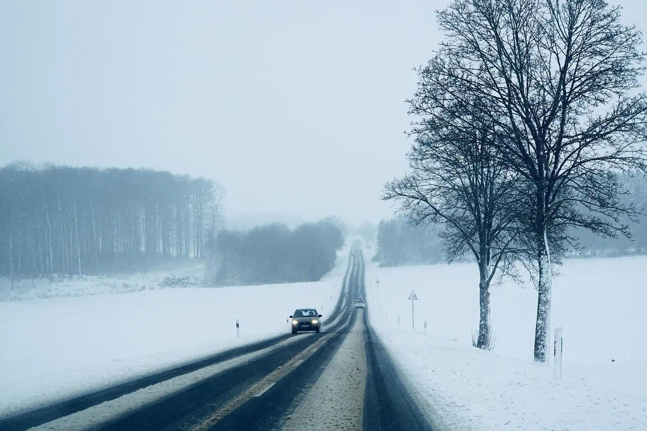 Route de campagne enneigée sous un ciel gris, une voiture phares allumés roule entre les champs blancs