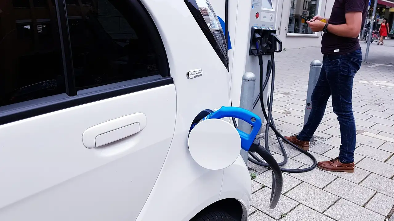 Petite voiture électrique blanche branchée à une borne de rue, un homme consulte son téléphone à côté