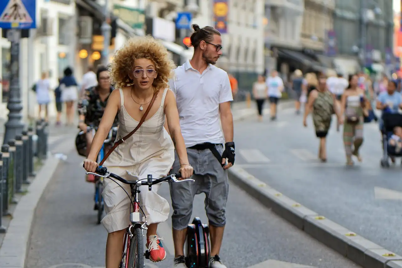 Une cycliste et un homme en gyroroue sur une piste cyclable, des piétons marchent sur la chaussée derrière eux