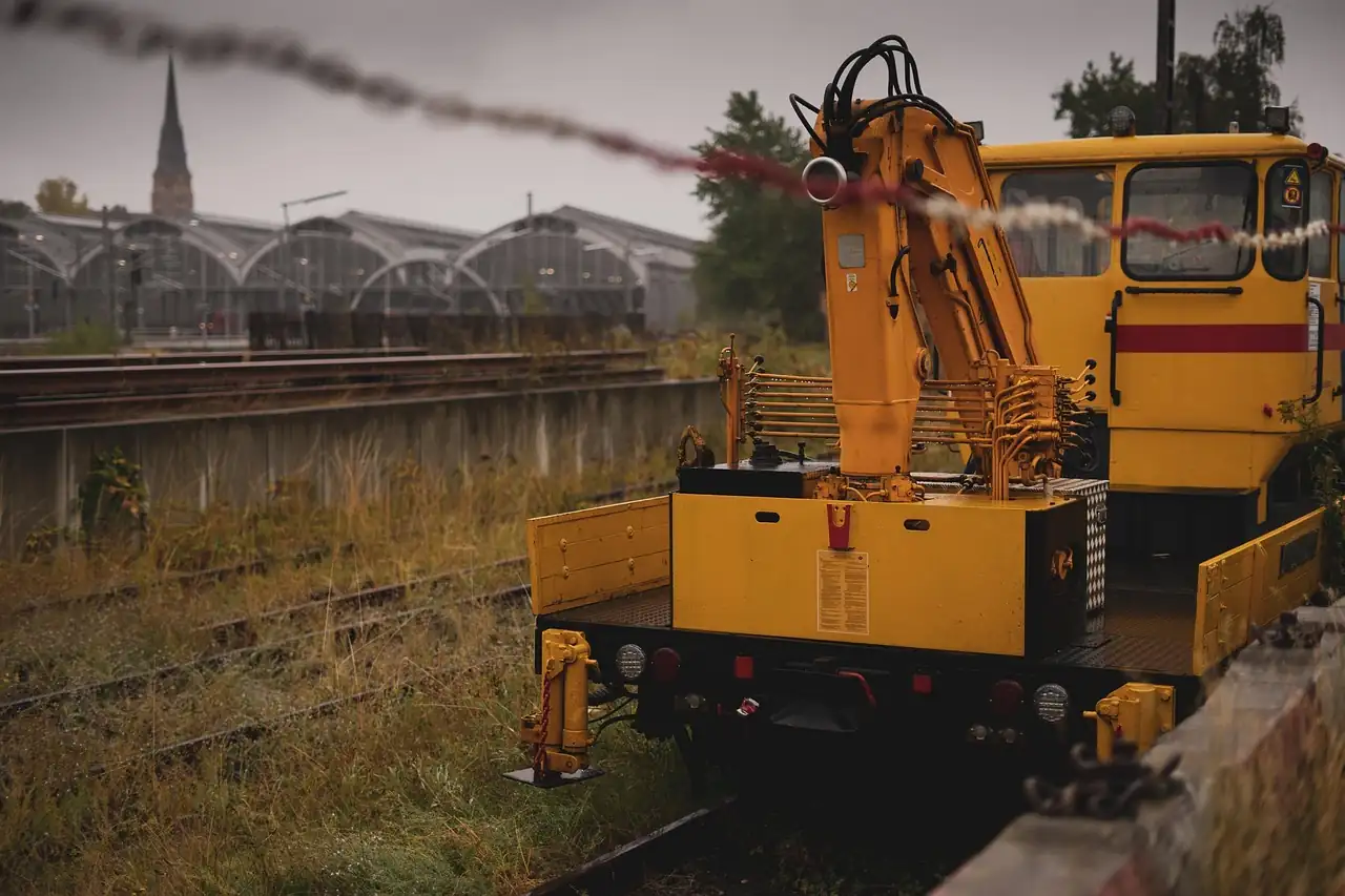 Engin de travaux jaune stationné sur une voie ferrée, grue repliée, sous un ciel gris de pluie