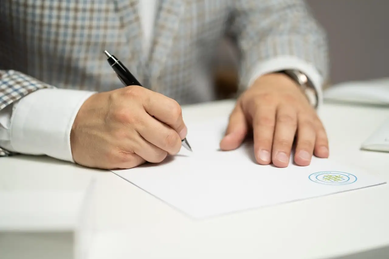 Un homme en veste à carreaux signe un document blanc posé sur un bureau, stylo noir à la main