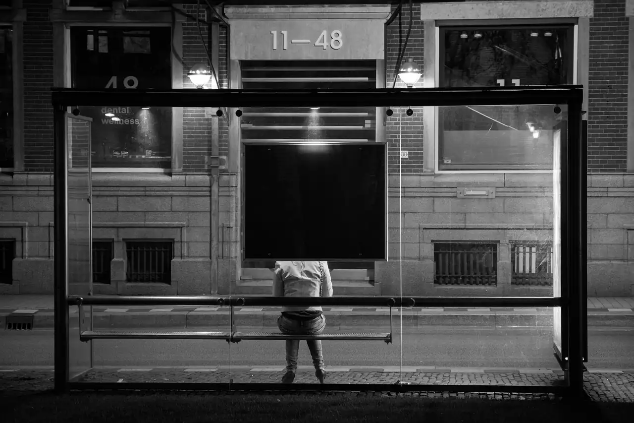 Personne seule assise sur le banc d'un abribus vitré la nuit, photo en noir et blanc, immeuble éclairé derrière