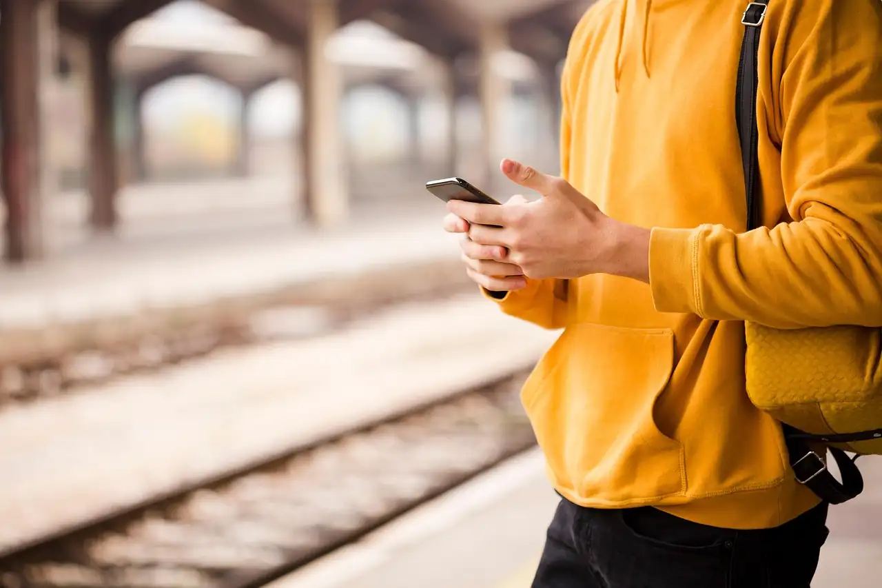 Personne en sweat jaune tenant un smartphone à deux mains sur un quai de gare flou, voie ferrée en contrebas