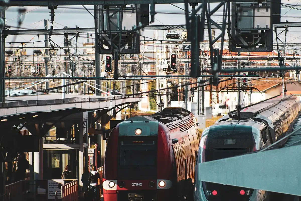 Deux rames SNCF, une rouge et une grise, arrêtées côte à côte à quai sous un enchevêtrement de caténaires et de signaux