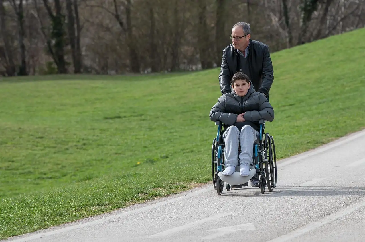 Homme poussant un enfant en fauteuil roulant sur une allée goudronnée le long d'une pelouse