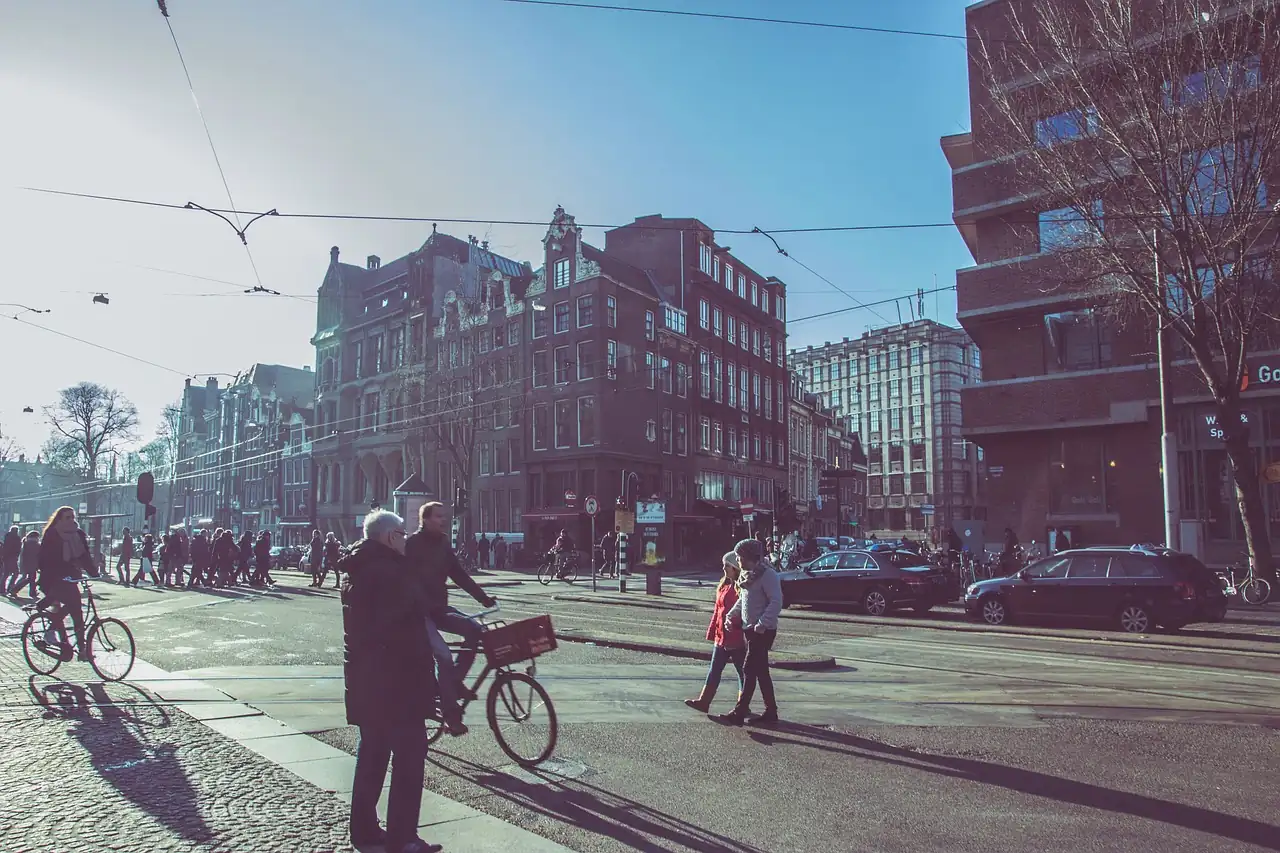 Cyclistes et piétons traversant un large carrefour d'Amsterdam en plein soleil, voitures à l'arrêt derrière