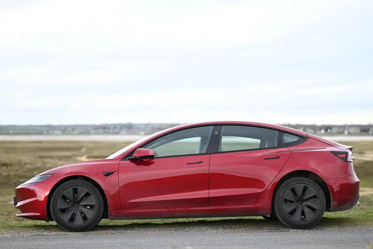 Berline électrique rouge garée de profil en bordure de prairie sous un ciel gris