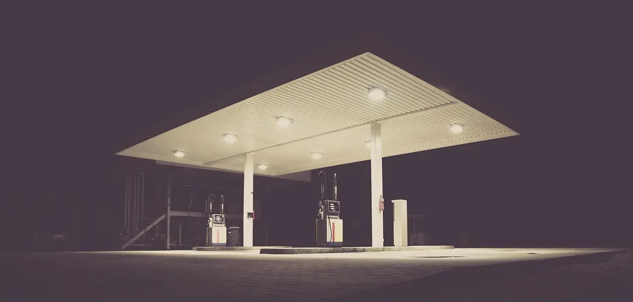 Station-service éclairée la nuit, auvent blanc au-dessus de deux pompes et rue déserte