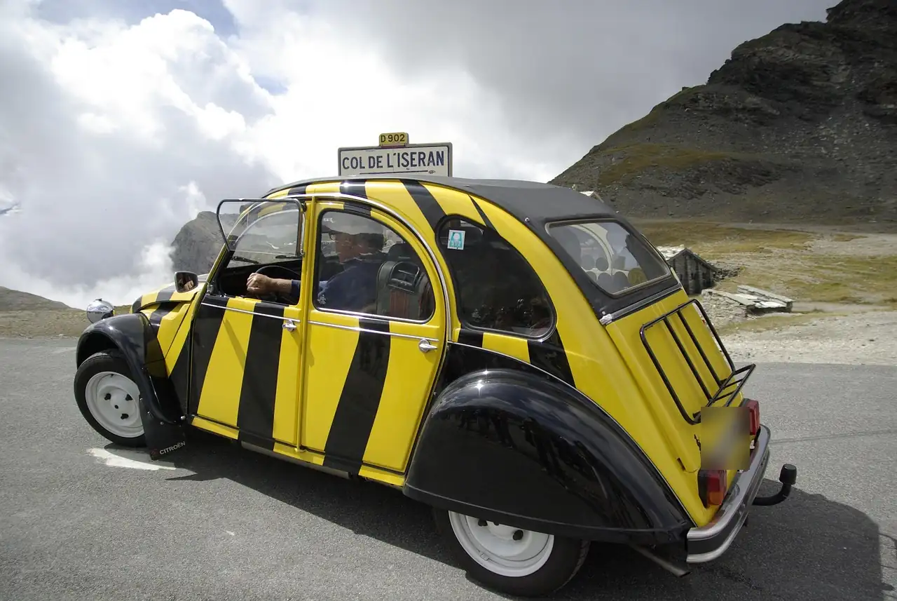 Citroën 2CV à rayures jaunes et noires garée en haute montagne devant un panneau de col routier, ciel nuageux
