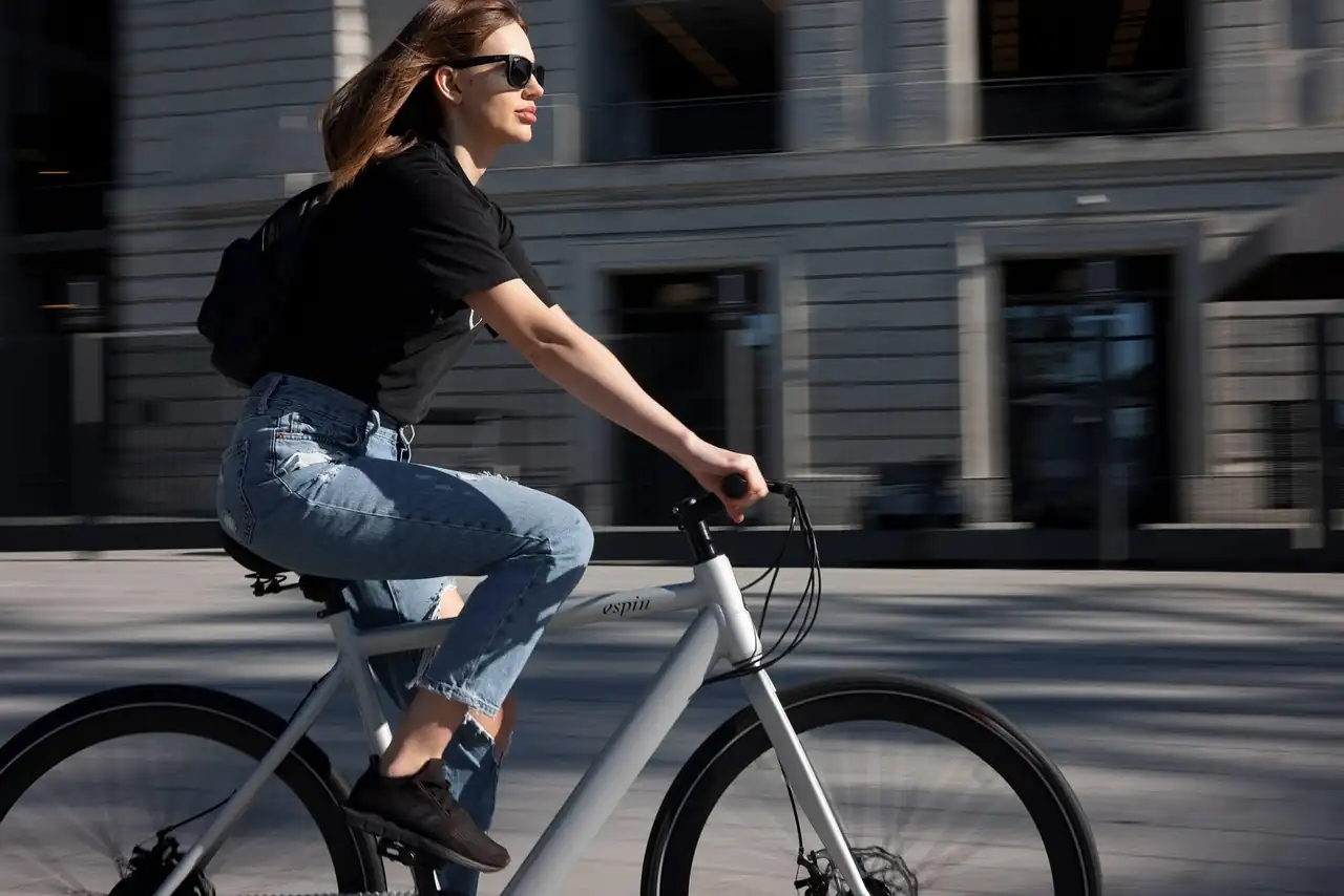 Jeune femme à lunettes de soleil roulant en ville sur un vélo blanc devant un immeuble gris